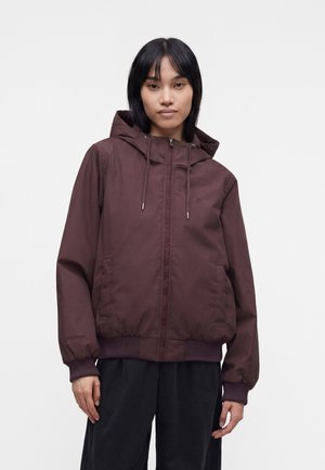 Femme portant une veste zippée à capuche marron foncé avec poignets et ourlet côtelés, debout devant un fond clair uni.