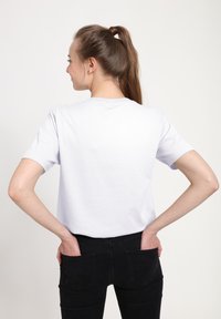 Bílé tričko s krátkým rukávem a kulatým výstřihem, zobrazené ze zad. V kombinaci s černými džíny, ruce resting v kapsách. Jednoduchý design.