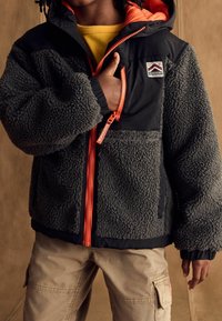 Child wearing dark gray fleece jacket with orange zipper and hood, yellow shirt underneath, and beige cargo pants, hand in chest pocket.
