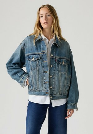 Kobieta z długimi blond włosami, ubrana w niebieską kurtkę jeansową na białą koszulę i ciemnoniebieskie jeansy, stojąca na jednolitym jasnym tle.