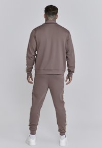 Chándal marrón hecho de un tejido suave con cuello alto, con franjas laterales de un color contrastante y puños ajustados, combinado con zapatillas blancas.