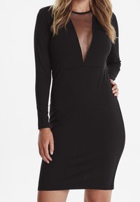 Femme portant une robe noire ajustée avec des manches longues et un décolleté en V transparent, se tenant avec une main sur la hanche.