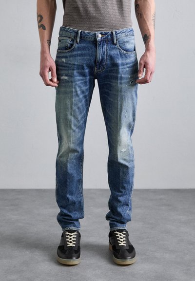 Jeans din denim de culoare albastru deschis, cu o ușoară decolorare și detalii uzate, asortate cu adidași negri cu șireturi albe și o talpă texturată.
