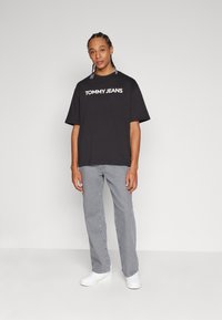 Camisa de tirantes negra con el logo "TOMMY JEANS" en blanco, combinada con jeans rectos gris claro y zapatillas blancas. Fondo sencillo.