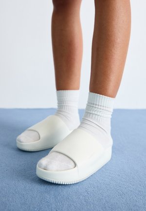 Jambes portant des chaussettes blanches et des sandales blanches à bouts ouverts, debout sur un sol moquetté bleu.