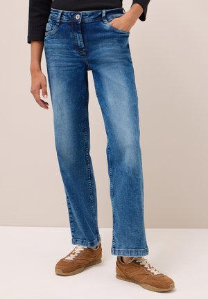 Persoon in blauwe vervaagde rechte jeans en bruine suède sneakers met witte veters, hand in zak tegen een neutrale achtergrond.