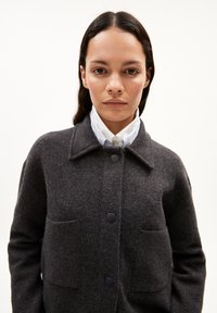 Giacca grigio scuro in misto lana con colletto, chiusura anteriore con bottoni e due tasche a toppa. Presenta una forma strutturata e una texture sottile.