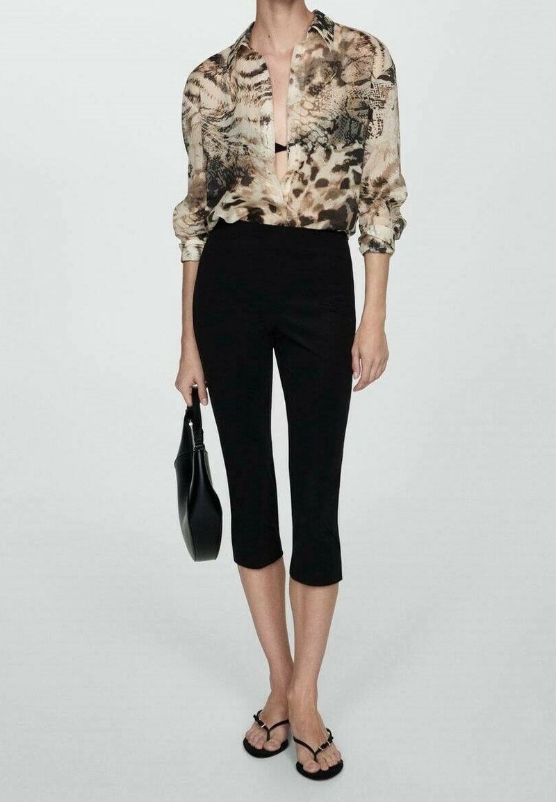 Personne portant un chemisier transparent à imprimé animal, un pantalon court noir, des sandales noires à talons, et tenant un sac à main noir sur un fond uni.