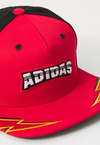 Raudona beisbolo kepurė su juodu viršumi, puošta ryškiu baltu "ADIDAS" logotipu ant pleistro. Geltoni žaibo akcentai ant krašto. Lygus paviršius.