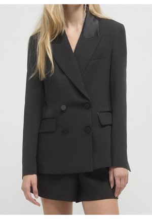 Woman wearing black double-breasted blazer with satin lapels and matching black shorts, standing against a plain light background.