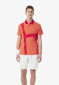Kortärmad polotröja i knallorange, med ett rött stänkmönster, ribbad krage och Lacoste-logotyp på bröstet. Bärs med vita shorts.