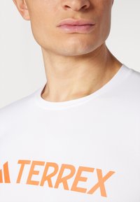 Valge tekstiilist särk, millel on ümmargune kaelus, sile tekstuur ja silmapaistev oranž "TERREX" logo ees. Suurendatud vaade ülemisele torsole.