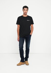 Sort bomulds t-shirt med et hvidt "REPLAY" logo på brystet, parret med mørke jeans og brune sneakers. Enkel, afslappet design.