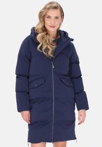 Femeie cu păr lung, ondulat și blond, îmbrăcată într-o geacă puffer de culoare bleumarin, cu lungimea până la genunchi, prevăzută cu fermoar și buzunare cu clapă, stând în fața unui fundal alb.