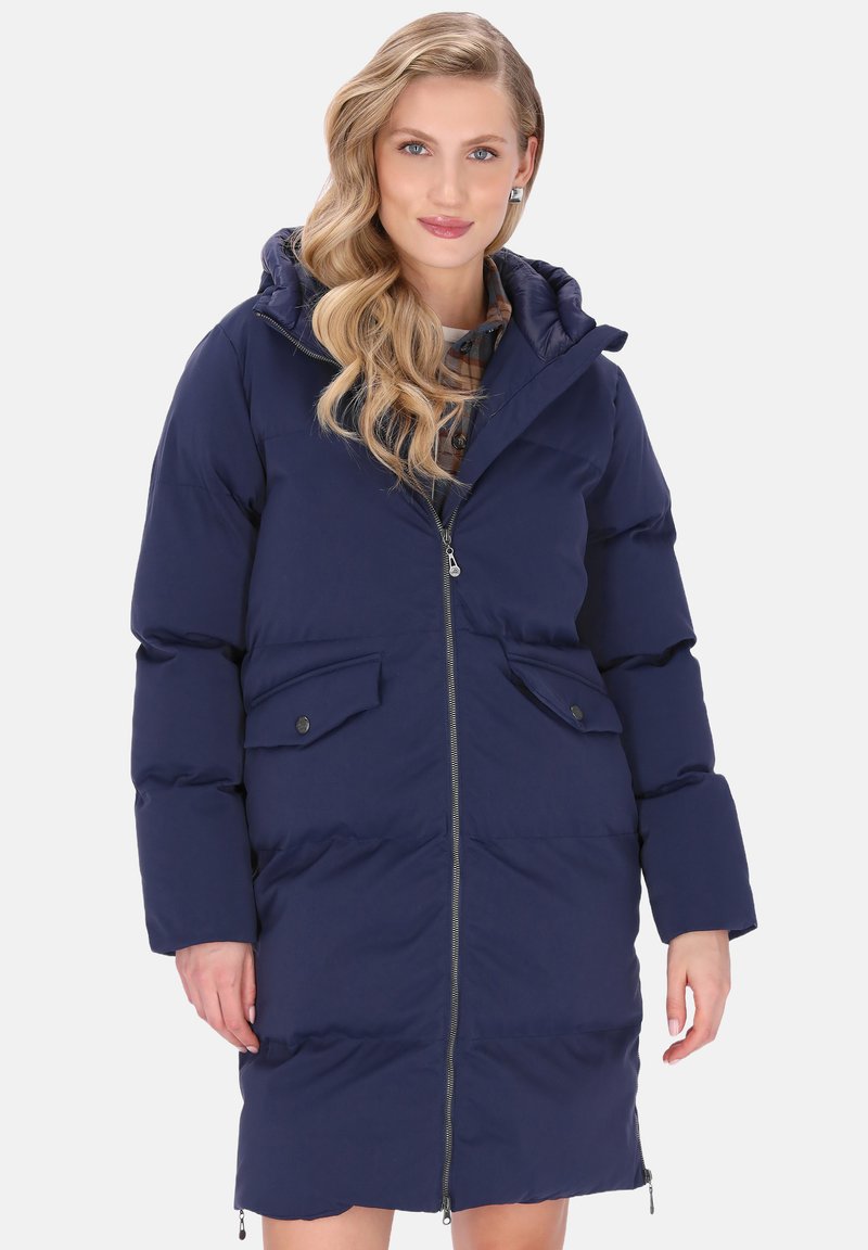 Femeie cu păr lung, ondulat și blond, îmbrăcată într-o geacă puffer de culoare bleumarin, cu lungimea până la genunchi, prevăzută cu fermoar și buzunare cu clapă, stând în fața unui fundal alb.