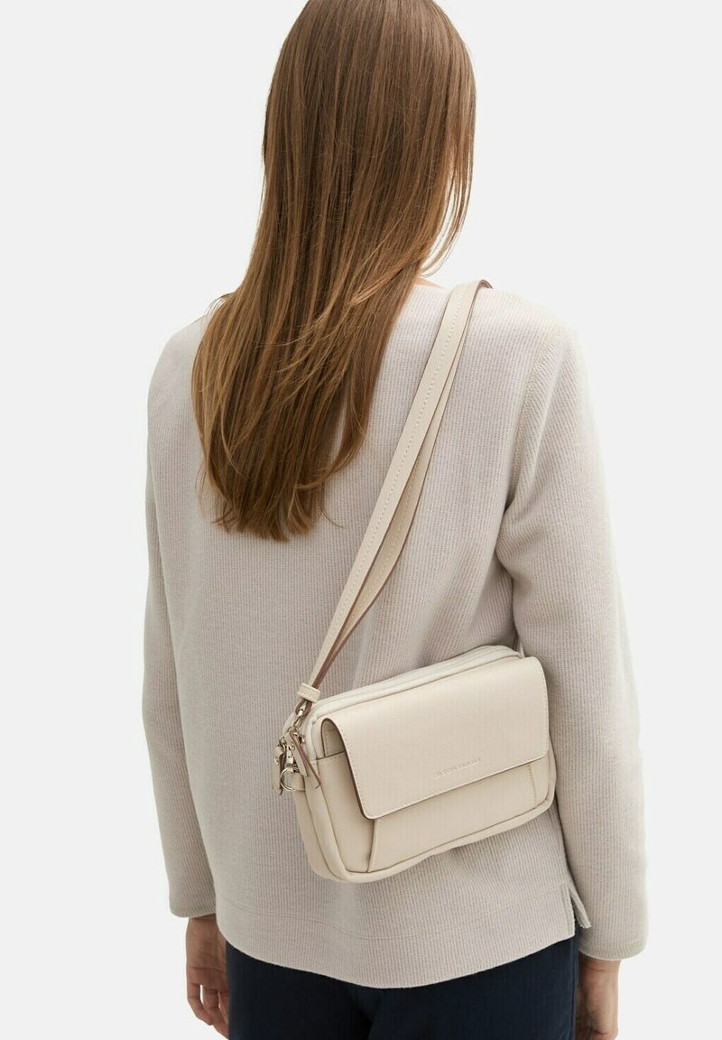 Beżowa torebka na ramię wykonana z teksturowanej skóry, z klapką na zapięcie i regulowanym paskiem, noszona na jasnym swetrze i ciemnych spodniach.