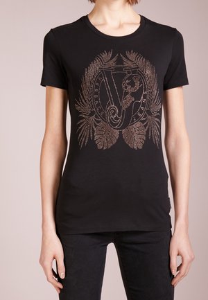 Kobieta nosząca czarny dopasowany t-shirt z ozdobnym metalowym emblematem przedstawiającym litery V i J otoczone wzorami liści.