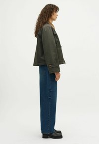 Olajzöld dzseki széles ujjal, nagy elülső zsebbel és bronz részletekkel, magas derekú kék farmerral és fekete bokacsizmával párosítva.