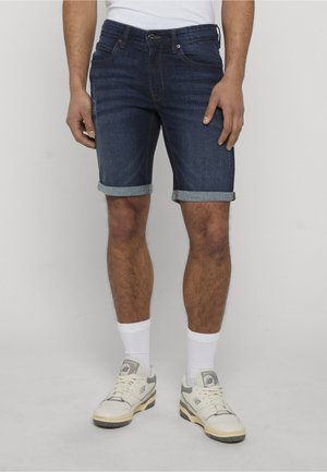 Homme portant un short en jean bleu foncé retroussé, des chaussettes blanches et des baskets blanches, debout devant un fond clair uni.