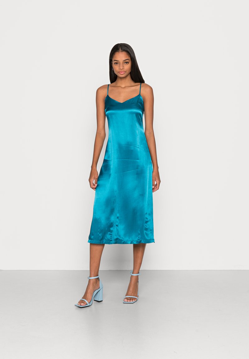 Vestido slip em cetim teal com alças finas, design em V e comprimento médio; combinado com sandálias de salto azuis claras.