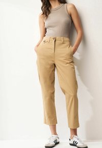 Kobieta w beżowej bezrękawnikowej bluzce, w wysokich khaki spodniankach z nogawkami 7/8 i białych trampkach z czarnymi paskami, stojąca z rękami w kieszeniach.