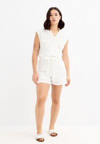 Mono corto blanco con detalles de ojal, diseño cruzado y mangas cortas. Presenta un cinturón atado en la cintura. Se lleva con sandalias de tiras blancas.