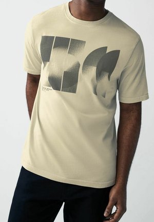 Homme portant un t-shirt beige à manches courtes avec des formes géométriques noires abstraites et un texte sur la poitrine, associé à un pantalon noir.