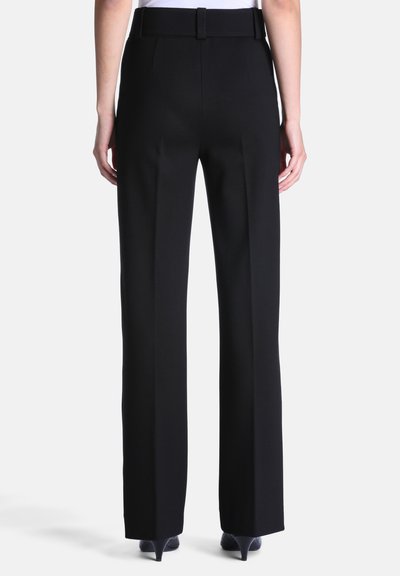 Personne portant un pantalon droit noir avec une ceinture assortie, des chaussures noires à talons, debout de dos sur un fond uni.