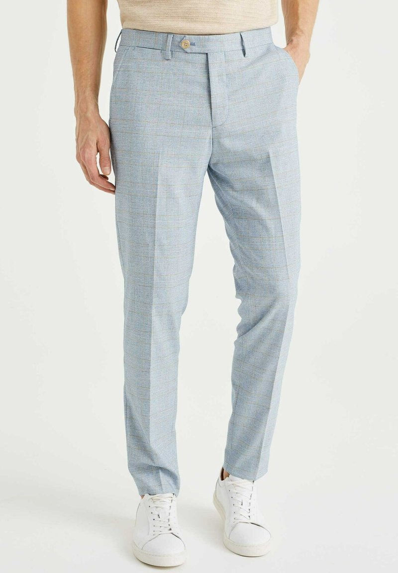 Muž oblečený v světle modrých kostkovaných slim-fit kalhotách a bílých teniskách, stojící s jednou rukou v kapse na jednobarevném pozadí.