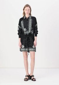 Svart skjortekjole med hvite mønstre, med snøring i midjen, lange ermer og en kontrasterende paisley-trykk langs kantene. Svart sandaler.