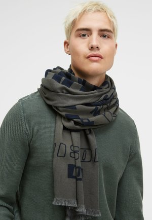 Gray scarf with navy geometric pattern, finished with fringed edges, worn over a textured green sweater. Soft fabric, lightweight design.