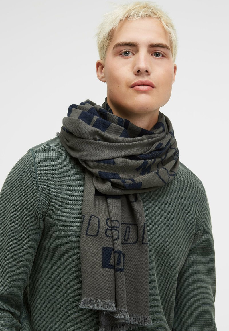 Gray scarf with navy geometric pattern, finished with fringed edges, worn over a textured green sweater. Soft fabric, lightweight design.