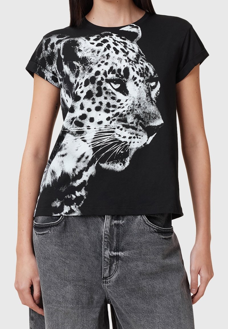 T-shirt noir à manches courtes avec un grand imprimé graphique de léopard blanc. Fabriqué en tissu doux avec une coupe décontractée, présenté avec un jean gris.
