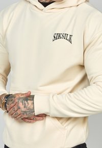 Persona vestida con una sudadera color crema de SikSilk con bolsillo tipo canguro, mostrando manos y muñecas tatuadas adornadas con pulseras.