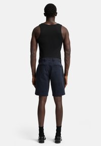 Shorts marine à la texture lisse, avec un devant plat et des poches arrière. Portés avec un débardeur noir et des chaussettes noires.