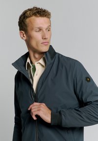 Marineblauwe jas met een hoge kraag, ritsluiting en geribbelde manchetten. Heeft een gestructureerde zwarte kraag en een logo-opdruk op de mouw.