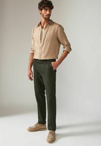 Kragskjorte i beige linned over en hvid T-shirt, parret med mørkegrønne bukser og brune sko med en struktureret overflade og snørebånd.