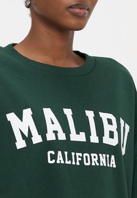 Grüner Pullover mit weißem Aufdruck "MALIBU" und "CALIFORNIA", geripptem Ausschnitt, weichem Stoff und lockerem Schnitt.
