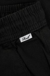Los pantalones elásticos negros presentan una cinturilla elástica con un acabado texturizado, acentuados por una pequeña etiqueta blanca con "Reell" en cursiva.