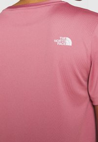 Camiseta de manga corta de color rosa claro, hecha de una tela suave y elástica. Presenta un pequeño logotipo blanco de The North Face en la parte superior de la espalda.