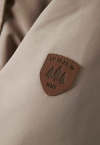 Beeziv kangas, millel on sile tekstuur ja pruun nahkplekk. Plekk on reljeefse kujundusega, kus on kujutatud kolme puud ja koordinaate.