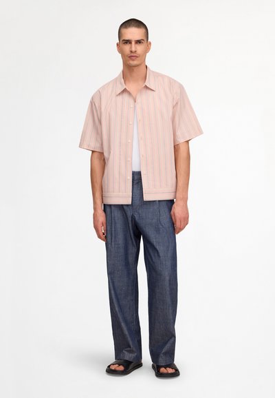 Homme portant une chemise à manches courtes rayée rose et blanche, un pantalon large bleu foncé, des sandales noires, debout devant un fond blanc.