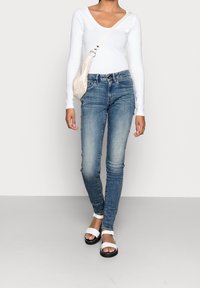 Top bianco a maniche lunghe, jeans blu aderenti e sandali bianchi e neri. I dettagli chiave includono uno scollo a barca e un lavaggio denim chiaro.