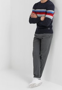 Pull marin bleu marine avec des rayures horizontales rouges, blanches et bleues, associé à un pantalon gris et des baskets blanches. Le matériau semble texturé et décontracté.