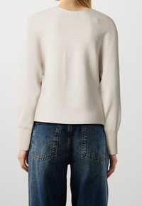 Pull beige clair en maille avec des côtes verticales, doté de manches longues et d'un col arrondi, associé à un jean large bleu foncé.