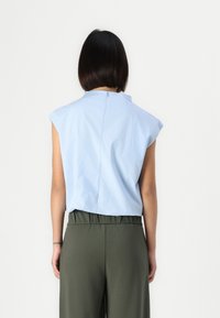 Top senza maniche azzurro chiaro con collo alto e cerniera sul retro, abbinato a pantaloni larghi color verde oliva. Tessuto dalla consistenza liscia.