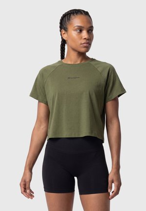 Woman with braided hair wearing an olive green cropped t-shirt and black high-waisted biker shorts, standing and looking to the side.