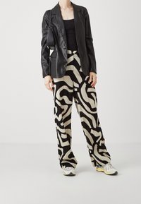 Zwart lederen blazer over een zwart kanten top, gecombineerd met beige wijde broek met een gedurfde zwarte spiraalpatroon en lichte sneakers.