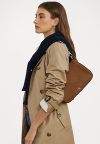 Brun läderhandväska med en slät yta, rektangulär form och guldfärgad hårdvara. Bärs på axeln tillsammans med en marinblå stickad scarf och en beige trench coat.