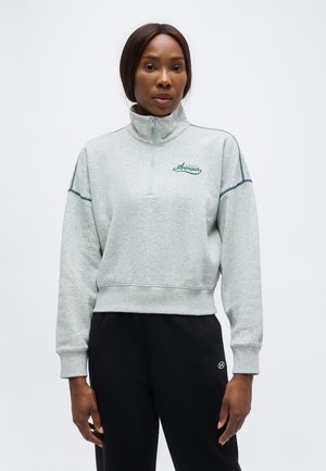 Kobieta w jasnoszarej bluzie z zamkiem do połowy, z zielonym logo "Under Armour" i czarnych spodniach, stojąca na jednolitym białym tle.